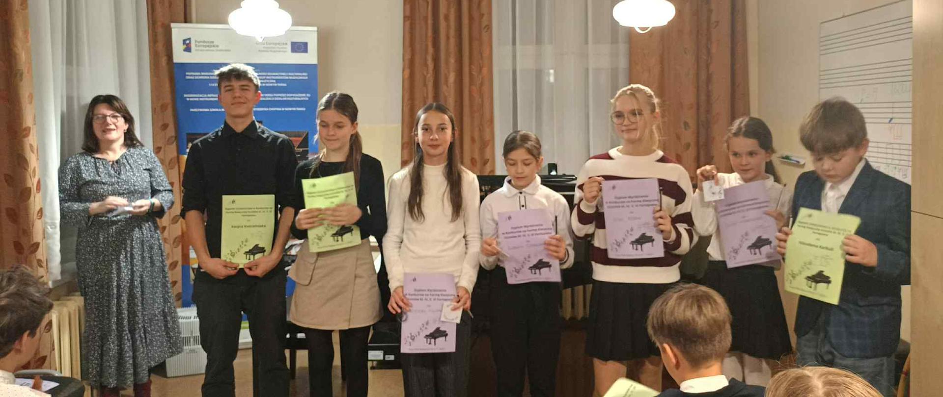 Zdjęcie przedstawia scenę z uroczystości wręczenia dyplomów po konkursie muzycznym. Akcja dzieje się w sali kameralnej. W tle widoczne są brązowe, ciężkie zasłony. Po lewej stronie wiszą plakaty informacyjne z logo instytucji, w tym logo Unii Europejskiej, sugerujące, że wydarzenie mogło być wspierane funduszami UE. Sufit oświetlają dwie wiszące lampy w stylu klasycznym, z jasnymi, szklanymi kloszami. Nauczycielka stoi po lewej stronie, ubrana jest w długą, wzorzystą sukienkę. Uśmiecha się i trzyma w rękach plik dyplomów, pełniąc rolę osoby wręczającej nagrody lub organizatorki.Na środku stoi sześcioro dzieci i młodzieży (trzech chłopców i trzy dziewczynki)Wszyscy trzymają w rękach dyplomy. Wysoki chłopiec, w czarnej koszuli i spodniach, stoi jako pierwszy. Uczennica w czarnym swetrze i beżowej spódnicy.
dziewczyna w białym, długim swetrze. Uczennica w białej bluzce, dalej uczennica w swetrze w biało-czerwone pasy i ciemnej spódniczce i na końcu chłopiec stoi na samym końcu, ubrany w marynarkę. Na dole zdjęcia, na pierwszym planie, widać fragmenty głów i pleców osób siedzących na widowni. Jeden chłopiec w granatowej marynarce odwrócony jest tyłem do obiektywu, obserwując scenę.
Ogólne Wrażenie: Zdjęcie uchwyciło uroczysty, ale radosny moment podsumowania jakiegoś osiągnięcia artystycznego, w atmosferze nagradzania młodego talentu.