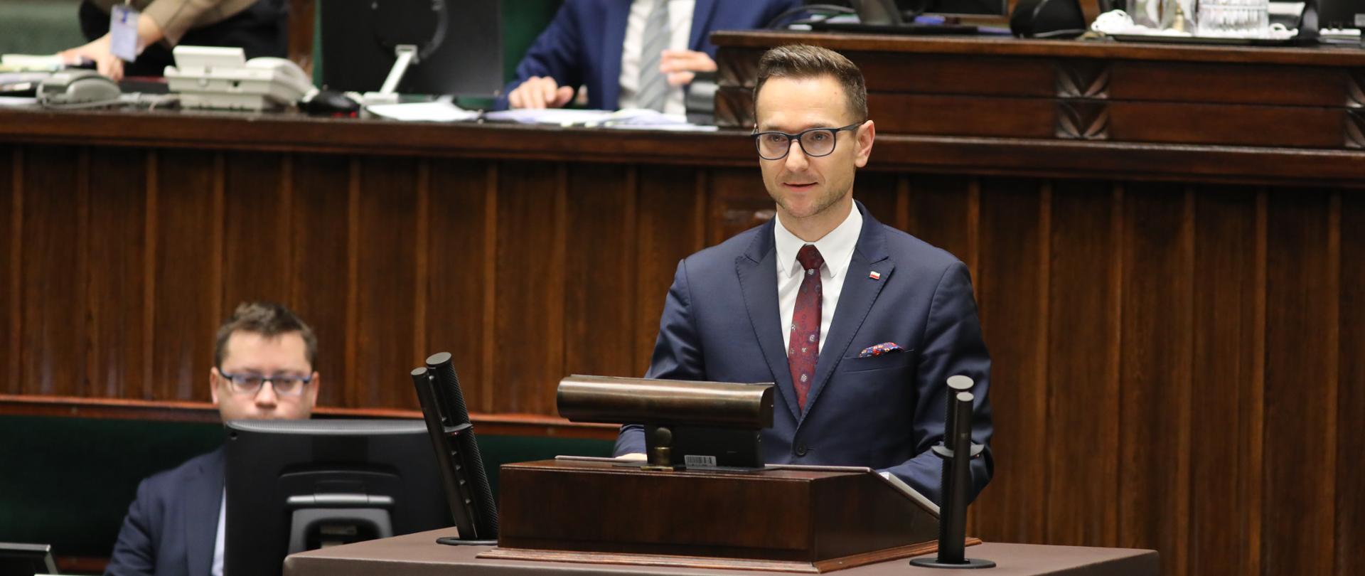 wiceminister Waldemar Buda stoi przed mównicą w sali plenarnej Sejmu i mówi do mikrofonu