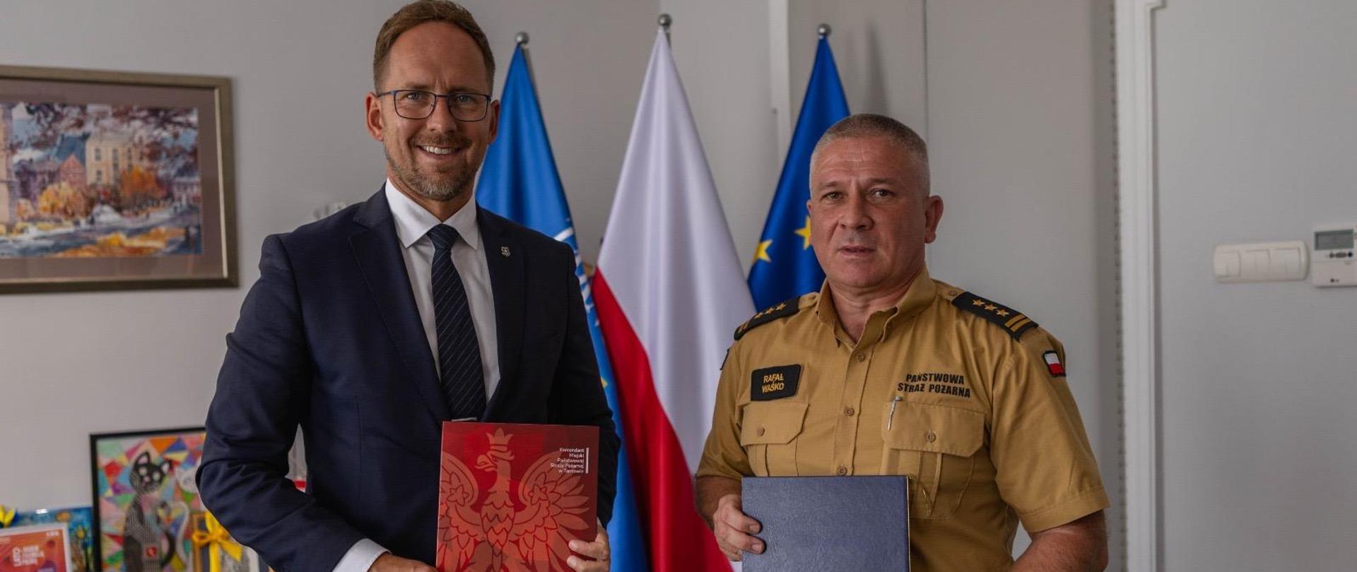 W pomieszczeniu z szarymi ścianami stoi prezydent miasta ubrany w granatowy garnitur oraz komendant w piaskowym mundurze. W rękach trzymają teczki z porozumieniem. W tle flaga Polski.