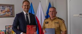 W pomieszczeniu z szarymi ścianami stoi prezydent miasta ubrany w granatowy garnitur oraz komendant w piaskowym mundurze. W rękach trzymają teczki z porozumieniem. W tle flaga Polski.