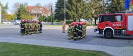Na placu przed Jednostka Ratowniczo Gaśnicza nr 1 stoją strażacy w dwóch szpalerach naprzeciw siebie na uroczystej zbiórce. Pomiędzy szpalerami stoi dowódca. Strażacy ubrani są w stroje bojowe. Za drugim szpalerem widoczny jest samochód gaśniczy.