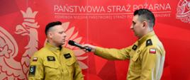 Na zdjęciu dwóch strażaków w beżowych mundurach stoi na tle czerwonej ścianki z napisem "Państwowa Straż Pożarna" oraz herbem. Jeden z nich trzyma mikrofon i przeprowadza wywiad z drugim, który patrzy w jego stronę. Obaj mają na mundurach oznaczenia stopni oraz polską flagę na ramieniu.