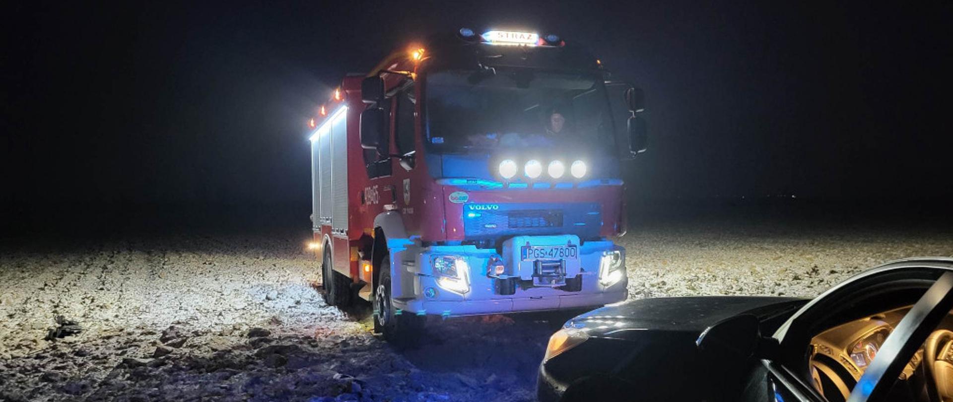 Samochód pożarniczy stojący na polu. Częściowo widać także wrak pojazdu osobowego po wypadku.