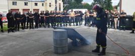 Warsztaty z zakresu fotowoltaiki. Plac Komendy Powiatowej PSP w Rawiczu, na którym odbywa się część praktyczna. Prowadzący prezentuje zachowanie się panelu fotowoltaicznego podczas działania na niego ogniem. Panel oparty jest o beczkę. W tle uczestnicy szkolenia oraz budynki komendy.