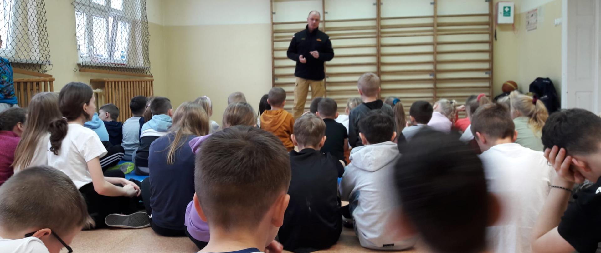 Na zdjęciu widać funkcjonariusza PSP ubranego w ubranie służbowe w trakcie pogadanki dla uczniów szkoły
podstawowej oraz przedszkola. Znajdują się na sali.