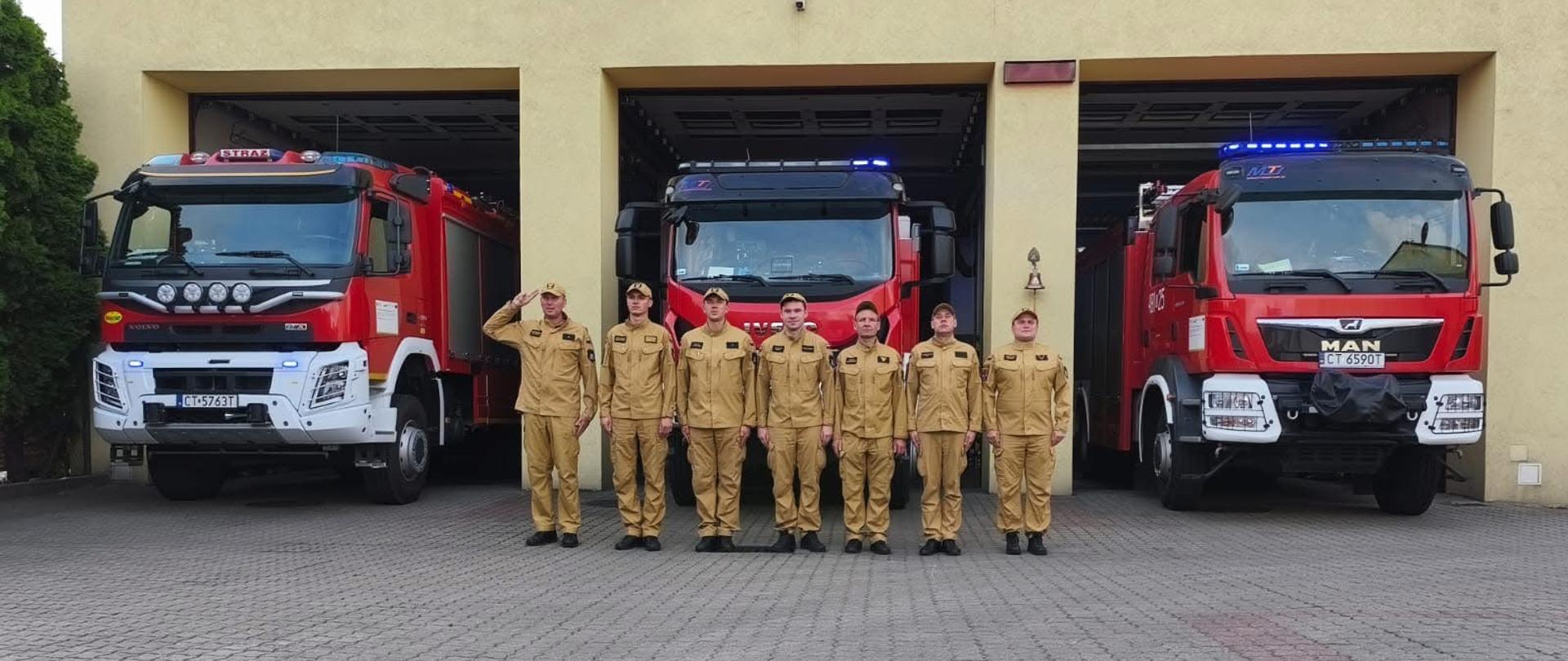 Strażacy stoją w szeregu i oddają hołd powstańcom warszawskim. W tle samochody pożarnicze z włączonymi sygnalizacjami świetlnymi. 