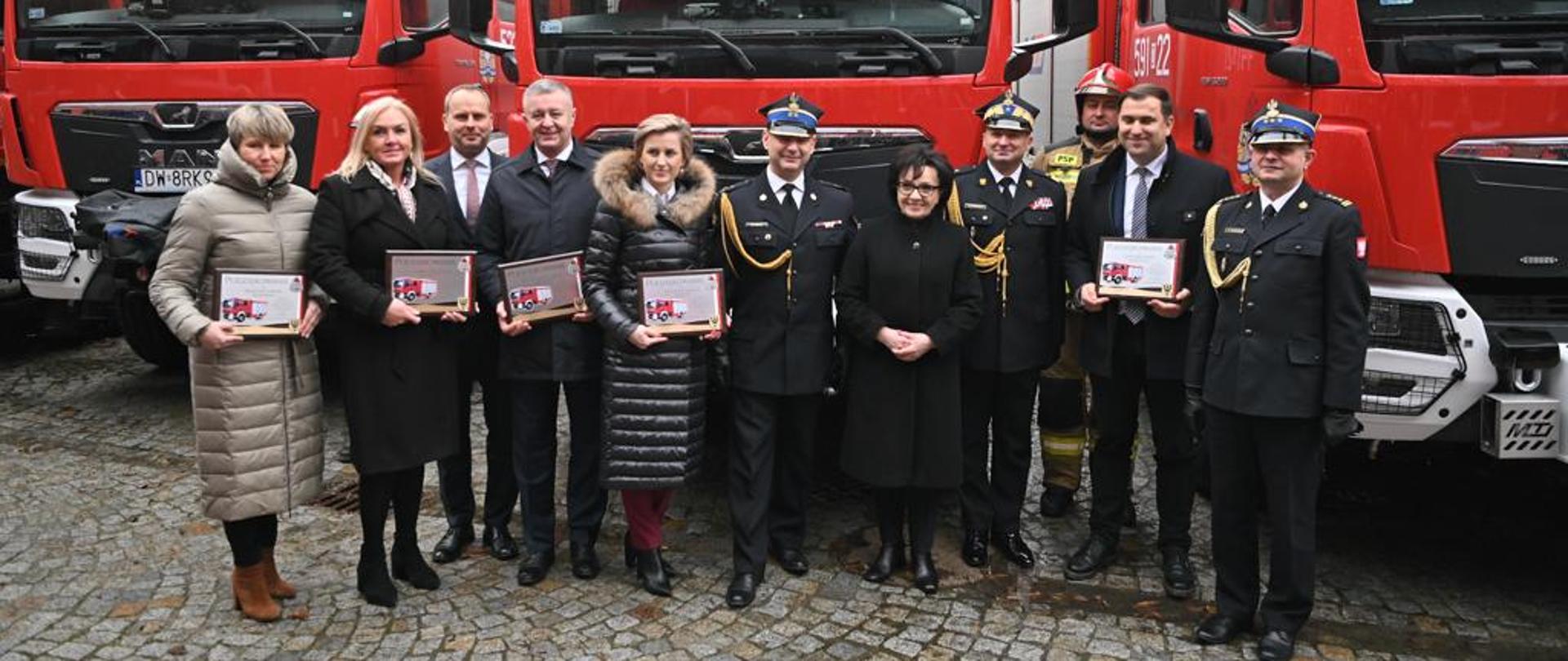 Obraz przedstawia Marszałek Sejmu, Zastępcę Komendanta Głównego, Komendanta Wojewódzkiego, Komendanta Powiatowego oraz przedstawicieli władz samorządowych z terenu powiatu jaworskiego na tle nowego samochodu ratowniczo-gaśniczego. Strażacy w umundurowaniu wyjściowym. Osoby znajdują sie na placu KW PSP we Wrocławiu.