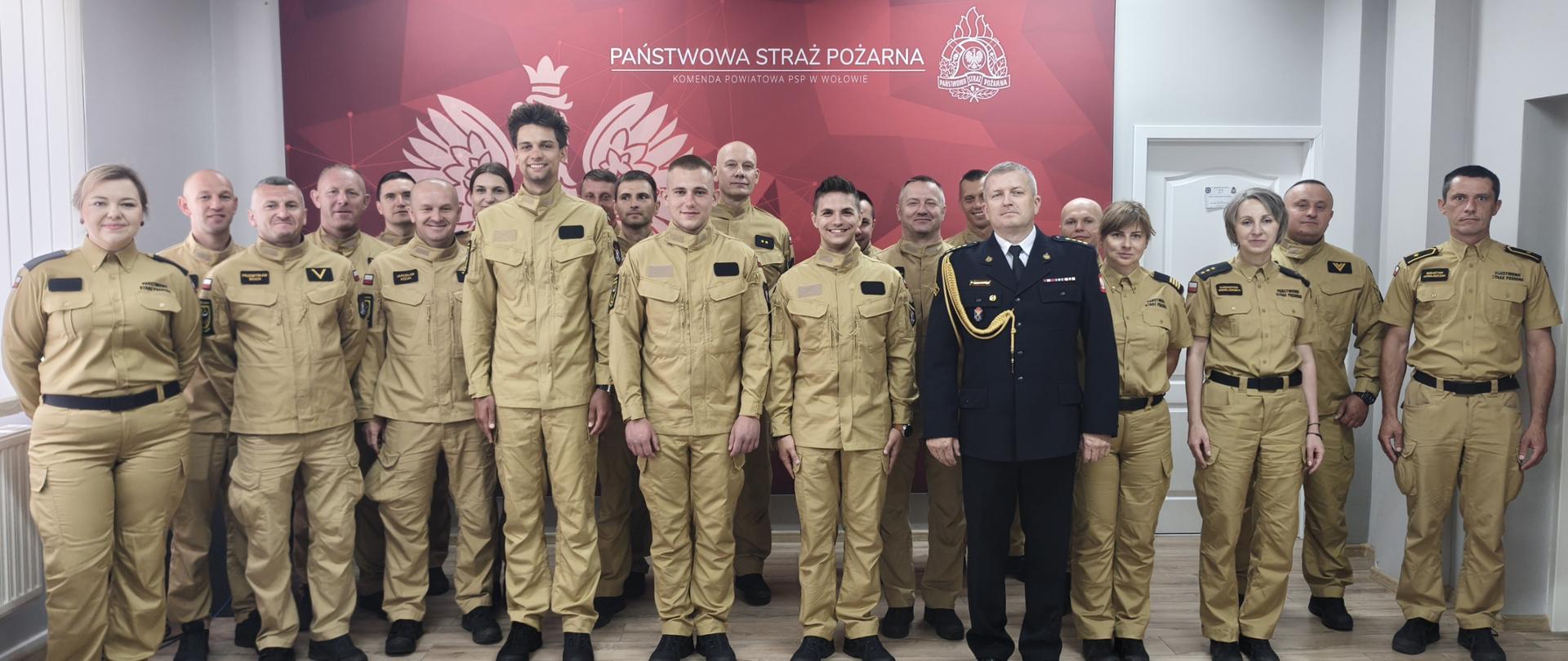 Pamiątkowe zdjęcie grupowe funkcjonariuszy Państwowej Straży Pożarnej w Wołowie. Duża grupa strażaków i strażaczek w piaskowych mundurach służbowych oraz jeden starszy oficer w ciemnym mundurze wyjściowym pozują wspólnie na tle czerwonej ścianki z godłem i napisem instytucji. Zdjęcie przedstawia oficjalne, pamiątkowe zdjęcie grupowe wykonane w pomieszczeniu z jasną, panelową podłogą. W tle widoczna jest duża, czerwona ścianka prezentacyjna z białym napisem „Państwowa Straż Pożarna Komenda Powiatowa psp w Wołowie” oraz grafiką przedstawiającą Orła Białego i emblemat strażacki. W kadrze znajduje się około 20 funkcjonariuszy (mężczyzn i kobiet), ustawionych w dwóch rzędach, którzy patrzą w stronę obiektywu i lekko się uśmiechają: Centrum pierwszego planu: Stoją trzej młodzi strażacy w piaskowych mundurach. Bezpośrednio obok nich, po prawej stronie, stoi starszy oficer w ciemnogranatowym mundurze wyjściowym ze złotym sznurem galowym na prawym ramieniu i baretkami odznaczeń. Resztę grupy tworzą strażacy i strażaczki ubrani w jednolite, piaskowe mundury służbowe. Część osób ma na sobie bluzy mundurowe, a część koszule. Wszyscy stoją w postawie wyprostowanej, co nadaje zdjęciu podniosły, a zarazem serdeczny charakter.