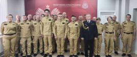 Pamiątkowe zdjęcie grupowe funkcjonariuszy Państwowej Straży Pożarnej w Wołowie. Duża grupa strażaków i strażaczek w piaskowych mundurach służbowych oraz jeden starszy oficer w ciemnym mundurze wyjściowym pozują wspólnie na tle czerwonej ścianki z godłem i napisem instytucji. Zdjęcie przedstawia oficjalne, pamiątkowe zdjęcie grupowe wykonane w pomieszczeniu z jasną, panelową podłogą. W tle widoczna jest duża, czerwona ścianka prezentacyjna z białym napisem „Państwowa Straż Pożarna Komenda Powiatowa psp w Wołowie” oraz grafiką przedstawiającą Orła Białego i emblemat strażacki. W kadrze znajduje się około 20 funkcjonariuszy (mężczyzn i kobiet), ustawionych w dwóch rzędach, którzy patrzą w stronę obiektywu i lekko się uśmiechają: Centrum pierwszego planu: Stoją trzej młodzi strażacy w piaskowych mundurach. Bezpośrednio obok nich, po prawej stronie, stoi starszy oficer w ciemnogranatowym mundurze wyjściowym ze złotym sznurem galowym na prawym ramieniu i baretkami odznaczeń. Resztę grupy tworzą strażacy i strażaczki ubrani w jednolite, piaskowe mundury służbowe. Część osób ma na sobie bluzy mundurowe, a część koszule. Wszyscy stoją w postawie wyprostowanej, co nadaje zdjęciu podniosły, a zarazem serdeczny charakter.