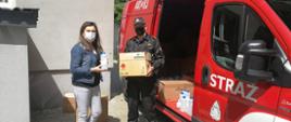Strażak wraz z dyrektorką placówki oświatowej w trakcie przekazania środków ochrony indywidualnej dla uczniów i pracowników szkoły, w tle samochód pożarniczy i budynek szkoły.