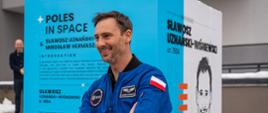 Sławosz Uznański-Wiśniewski w niebieskiej kurtce stoi na tle biało-niebieskiego słupa z napisem Poles in space.