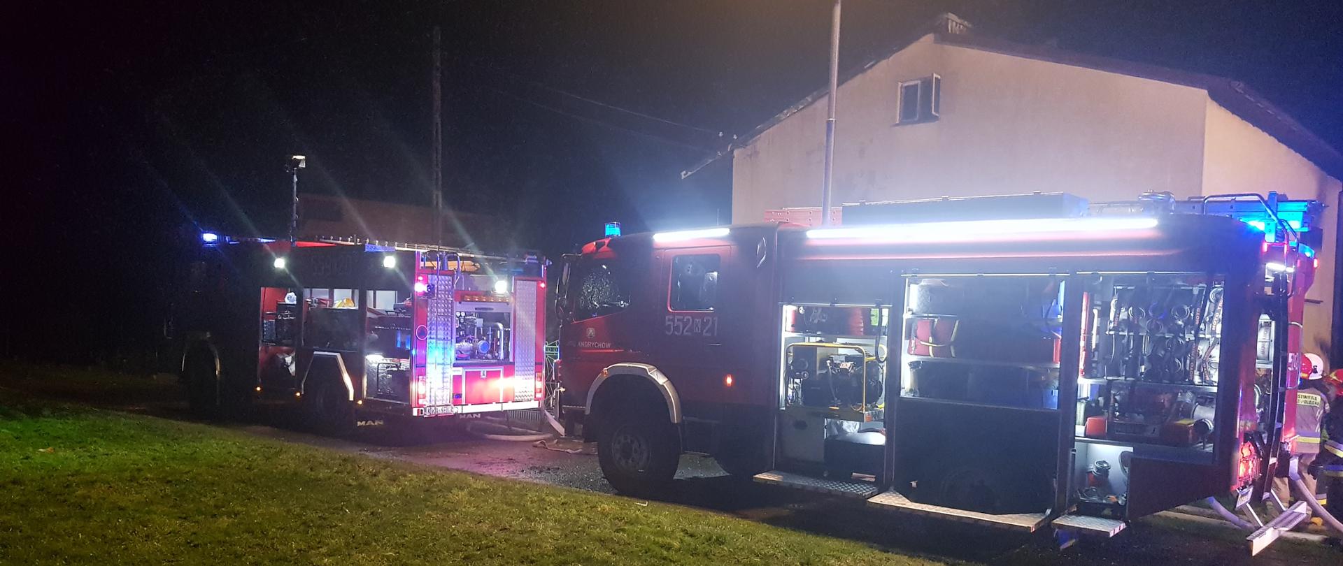 Widok nocą z ulicy na dwa samochody strażackie. Oba mają otwarte skrytki na sprzęt i rozstawione maszty oświetleniowe. Za samochodami budynek mieszkalny.