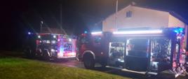 Widok nocą z ulicy na dwa samochody strażackie. Oba mają otwarte skrytki na sprzęt i rozstawione maszty oświetleniowe. Za samochodami budynek mieszkalny.