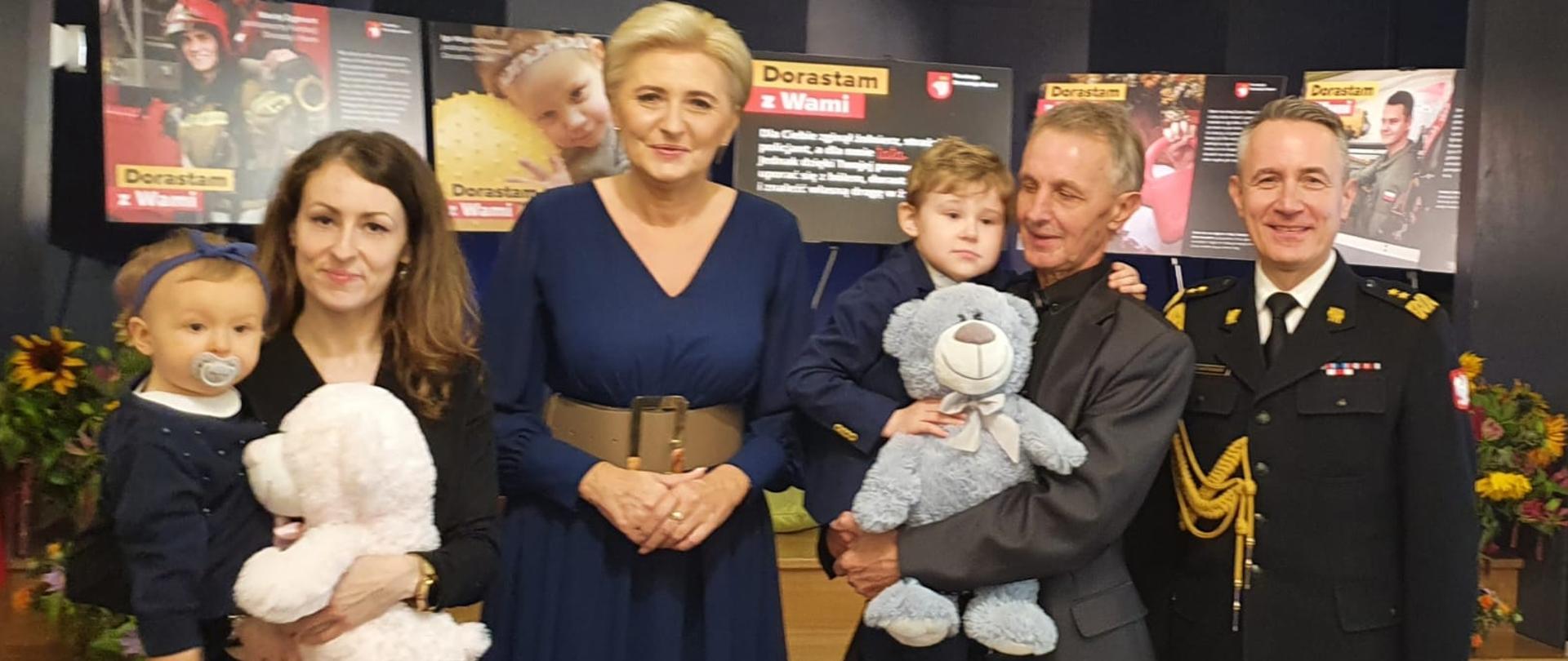 Na zdjęciu znajduje się Małżonka Prezydenta RP Agata Kornhauser-Duda razem z komendantem głównym PSP oraz rodziną zmarłego strażaka pozują do zdjęcia. W tle banery Fundacji 