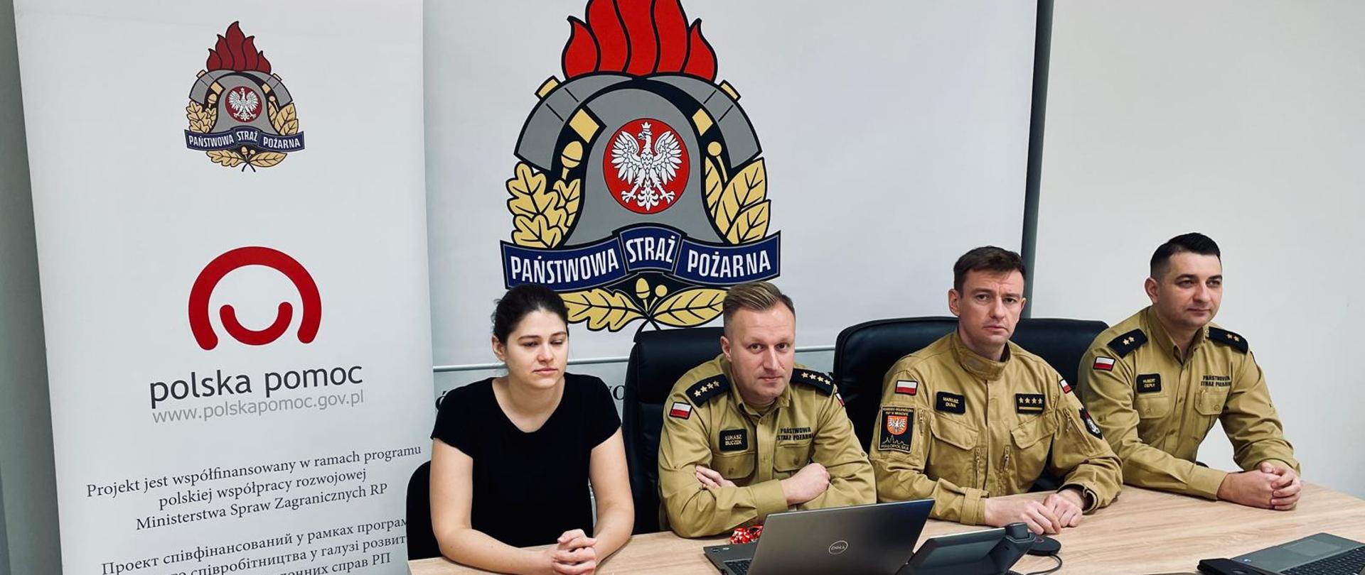 3 mężczyzn w mundurach i kobieta siedzą przygotowując się do zajęć online. Z tyłu baner z logo Polskiej pomocy i Państwowej Straży Pożarnej
