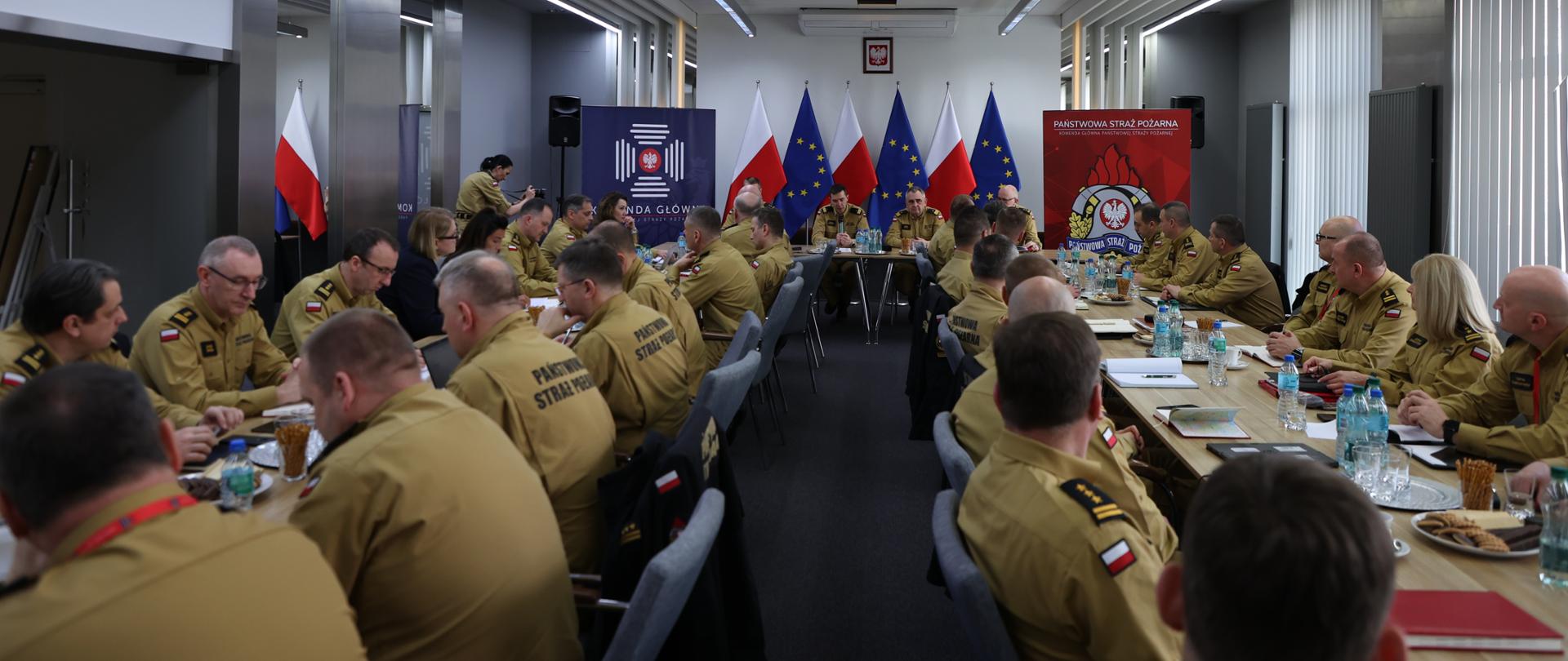 Kadra kierownicza zajęła miejsca siedzące podczas narady. Funkcjonariusze Państwowej Straży Pożarnej w mundurach służbowych uczestniczą w naradzie. W tle widoczne kierownictwo Komendy Głównej Państwowej Straży Pożarnej oraz banery KG PSP oraz flagi Polski i Unii Europejskiej.