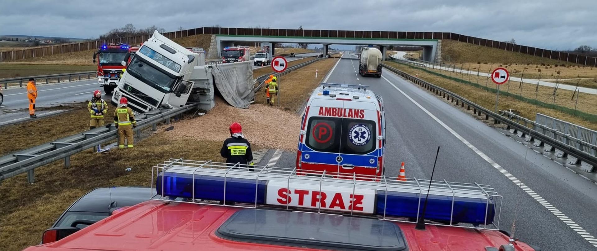 Zdjęcie zrobione z dachu samochodu ratowniczo-gaśniczego, na którym widnieją białe numery operacyjne 419-07. Przed pojazdem widać przewróconą na lewy bok ciężarówkę, z której wysypało się kruszywo. Przed pojazdem pożarniczym zaparkowana na jezdni karetka pogotowia. Na jezdni po lewej stronie strażacy w umundurowaniu specjalnym, którzy zajmują się techniczna stroną zdarzenia.