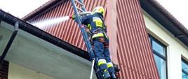 Na zdjęciu widocznych czterech strażaków zabezpieczających drabinę pożarniczą w ubraniach specjalnych koloru piaskowego i granatowego w białych hełmach oraz jeden strażak na drabinie podający prąd wody na dach budynku. Budynek KP PSP elewacja biała dach blacha trapezowa koloru brązowego.