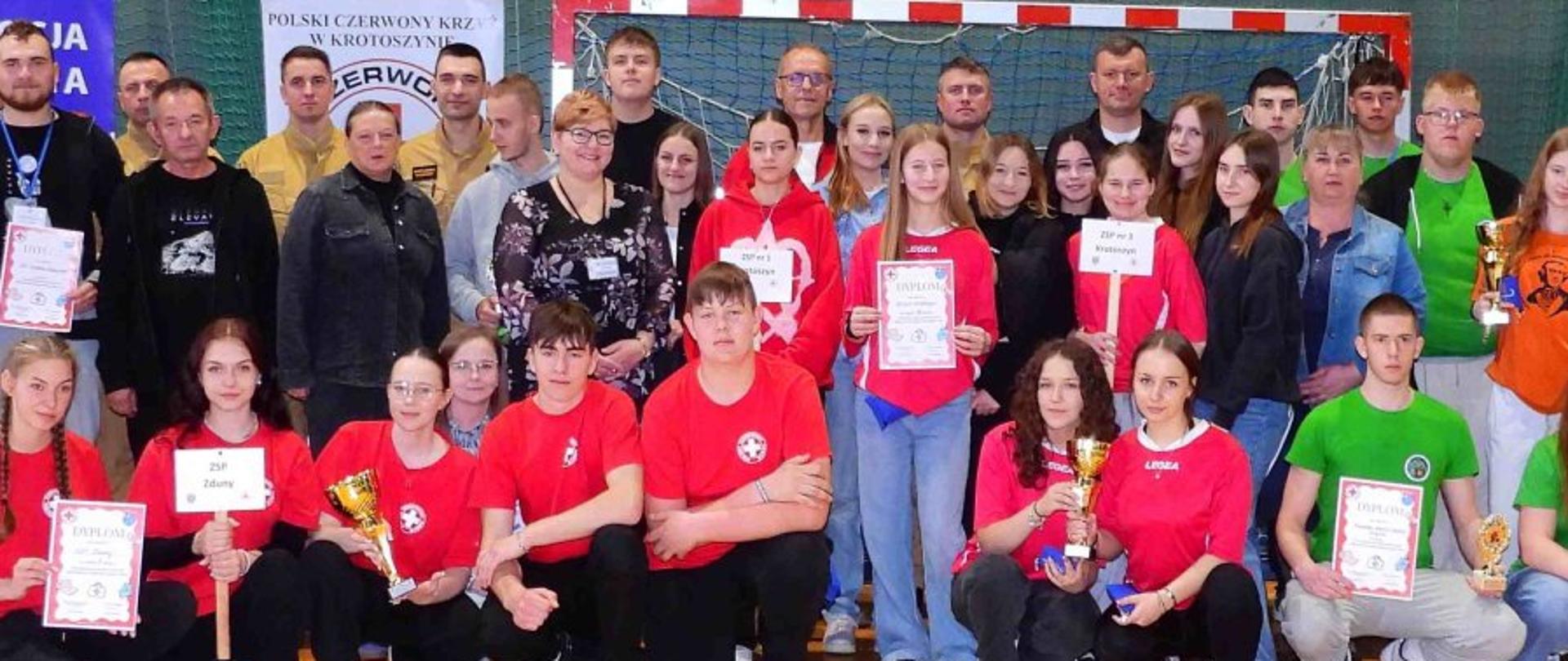Na zdjęciu widać dużą grupę ludzi zgromadzonych w hali sportowej. Na pierwszym planie siedzi i klęczy młodzież w czerwonych koszulkach z logotypem przypominającym symbol Polskiego Czerwonego Krzyża – część z nich trzyma puchary i dyplomy. W tle stoją dorośli – prawdopodobnie organizatorzy lub opiekunowie – oraz kolejni uczestnicy w różnokolorowych ubraniach (zielonych, czarnych, pomarańczowych). W tle widoczna jest bramka do piłki ręcznej oraz banery Polskiego Czerwonego Krzyża. Atmosfera na zdjęciu wydaje się radosna i dumna – wygląda na podsumowanie zawodów lub konkursu związanego z pierwszą pomocą.