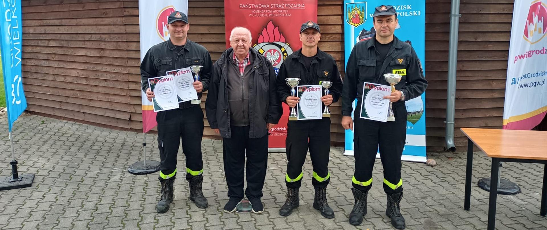Zdjęcie przedstawia trzech strażaków z nagrodami i kierownika drużyny stojących na tle drewnianego wigwamu