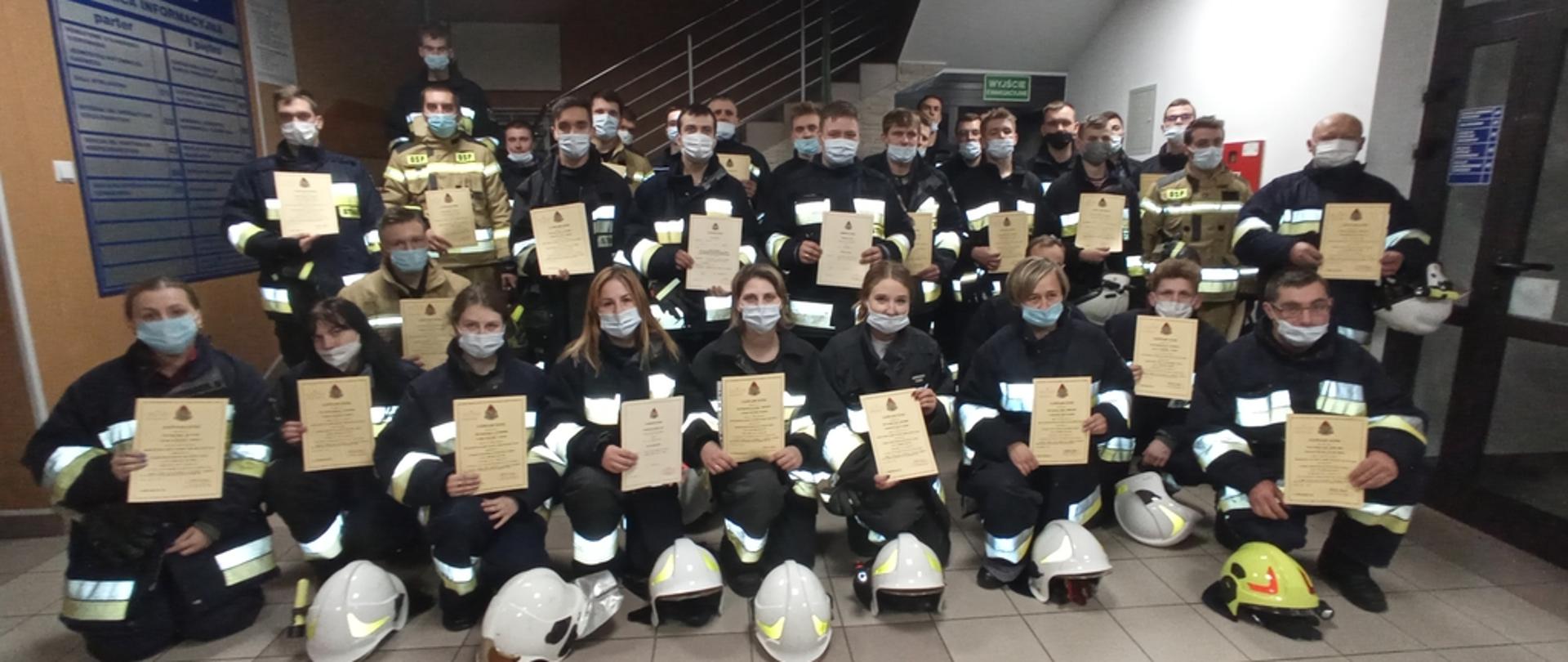 Na zdjęciu strażacy z Ochotniczych Straży Pożarnych po egzaminie kończącym szkolenie podstawowe strażaków ratowników OSP. Strażacy ubrani w ubrania specjalne koloru czarnego bez czapek trzymający w rękach zaświadczenia ukończenia szkolenie. 