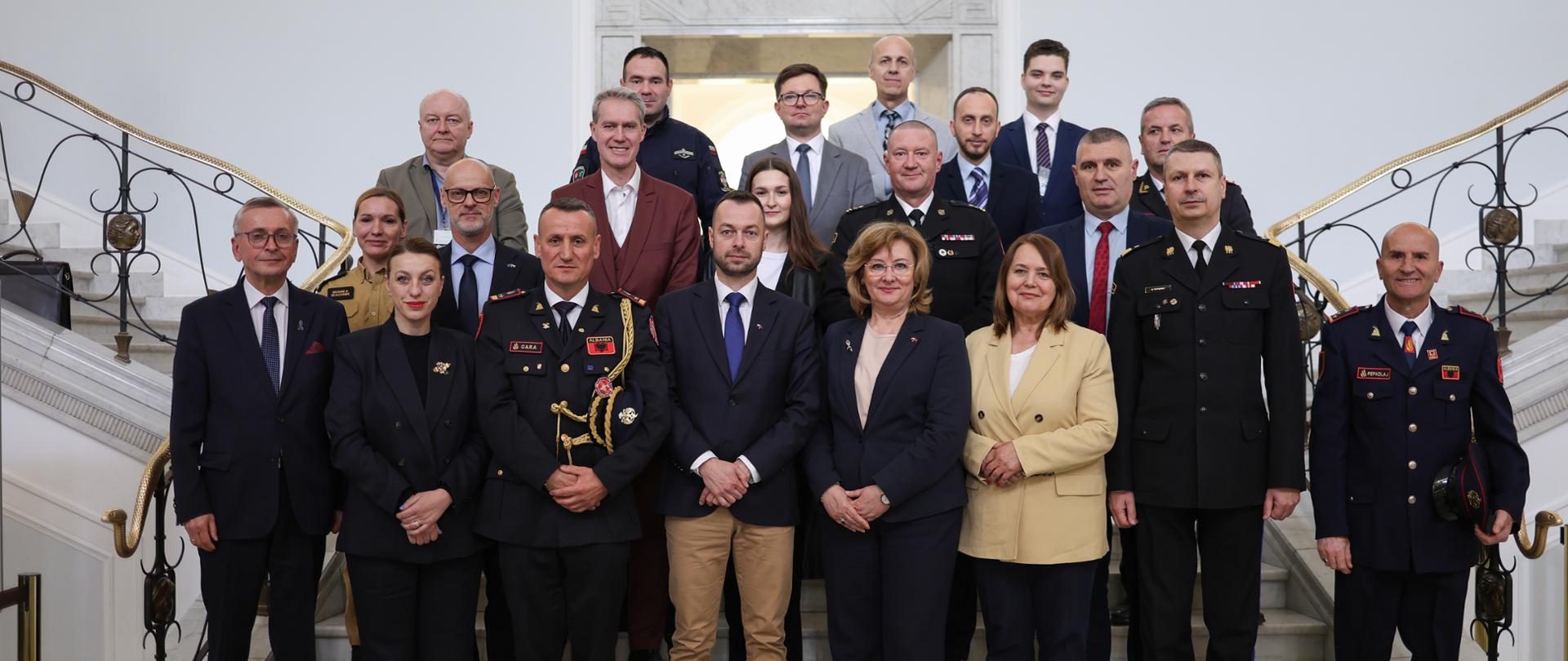 Delegacja Albanii oraz przedstawiciele PSP i administracji publicznej pozują do wspólnego zdjęcia na schodach w budynku instytucji państwowej.