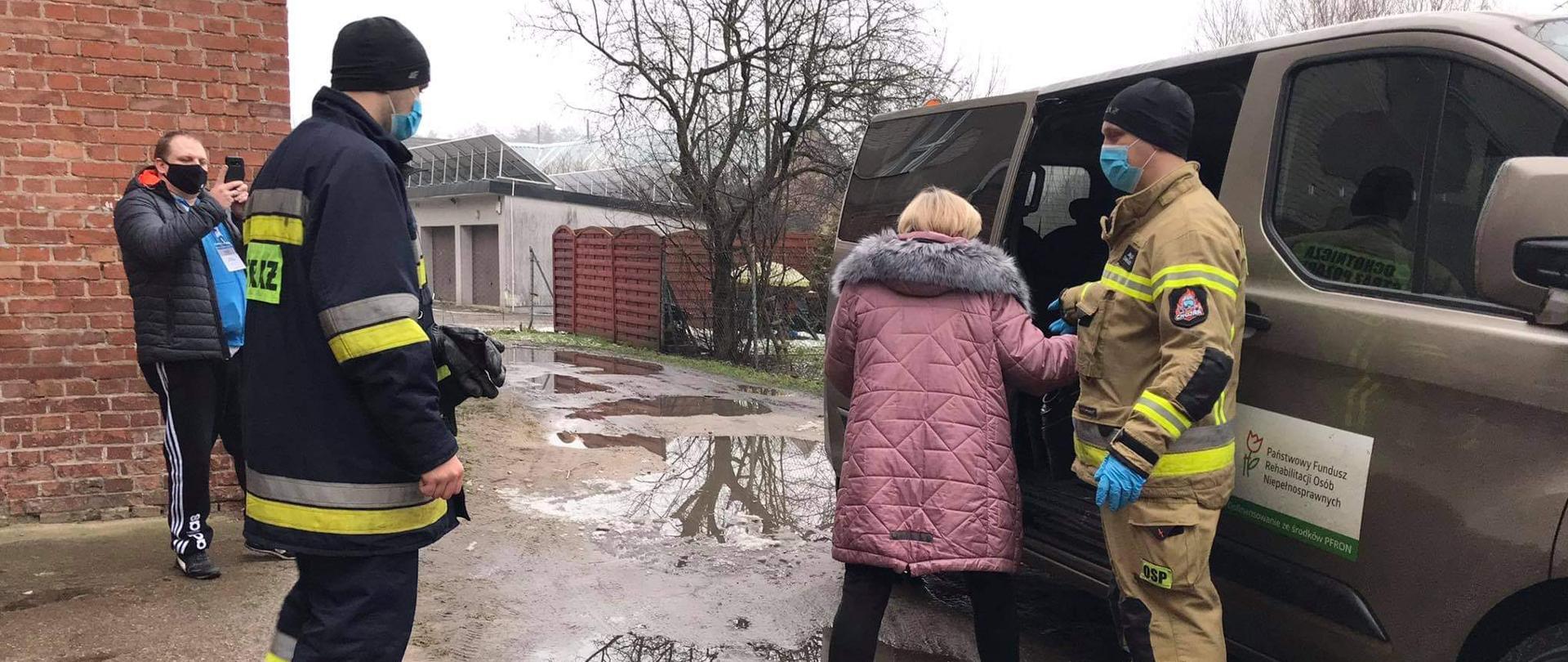 Zdjęcie przedstawia strażaków, którzy pomagają Pani wsiąść do samochodu. W tle kierowca samochodu robiący zdjęcie.