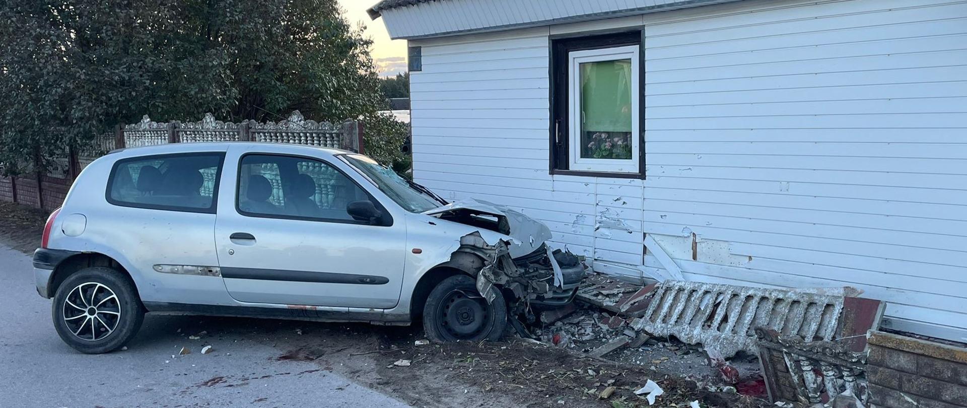 Zdjęcie przedstawia srebrny samochód osobowy, który uderzył w ścianę białego, parterowego domu, powodując znaczne uszkodzenia zarówno pojazdu, jak i budynku. Samochód ma mocno zniszczony przód, wgnieciony zderzak i częściowo oderwane koło. Ściana domu, obita białym sidingiem, jest wgnieciona i uszkodzona w miejscu uderzenia, a elementy tynkowe lub drewniane są oderwane. Widoczne są również zniszczone części, które mogły być fragmentem ogrodzenia lub dekoracji przed domem, leżące na ziemi. Obok miejsca zderzenia, pod oknem, znajduje się zniszczony element przypominający ogrodzenie lub ławkę. Po prawej stronie zdjęcia widoczne są ciemne drzwi wejściowe do domu, a nad nimi, po prawej stronie, kawałek zadaszenia. Po lewej stronie kadru widoczne są drzewa i krzewy. Światło na zdjęciu sugeruje poranek lub późne popołudnie. Cała scena wskazuje na poważny wypadek drogowy z kolizją z budynkiem.