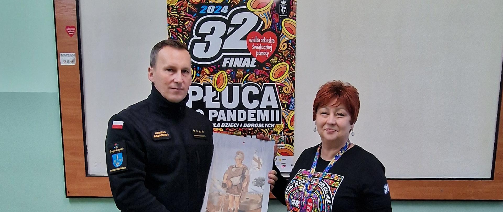 Strażak przekazujący płytkę z wizerunkiem św. Floriana wolontariuszowi WOŚP, w tle plakat 32 finału WOŚP