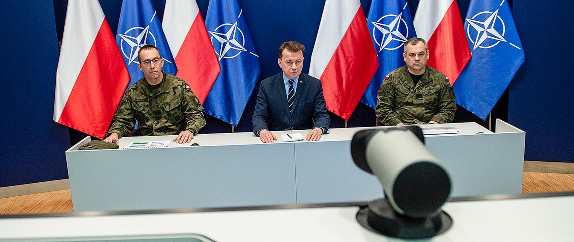 Na zdjęciu widoczny Mariusz Błaszczak, minister obrony narodowej w środku przy biurku. Po lewej stronie generał broni Tomasz Piotrowski, Dowódca Operacyjny, po prawej generał dywizji Wisław Kukuła, Dowódca Wojsk Obrony Terytorialnej. W kadrze widoczny monitor i kamera. W tle flagi Polski i Sojuszu Północnoatlantyckiego.