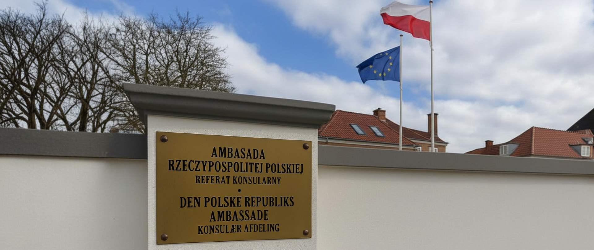 Tablica z napisem ‘Ambasada Rzeczypospolitej Polskiej – Referat Konsularny’ umieszczona na ogrodzeniu budynku ambasady; w tle widoczne flagi Polski i Unii Europejskiej na masztach oraz fragmenty okolicznych zabudowań.