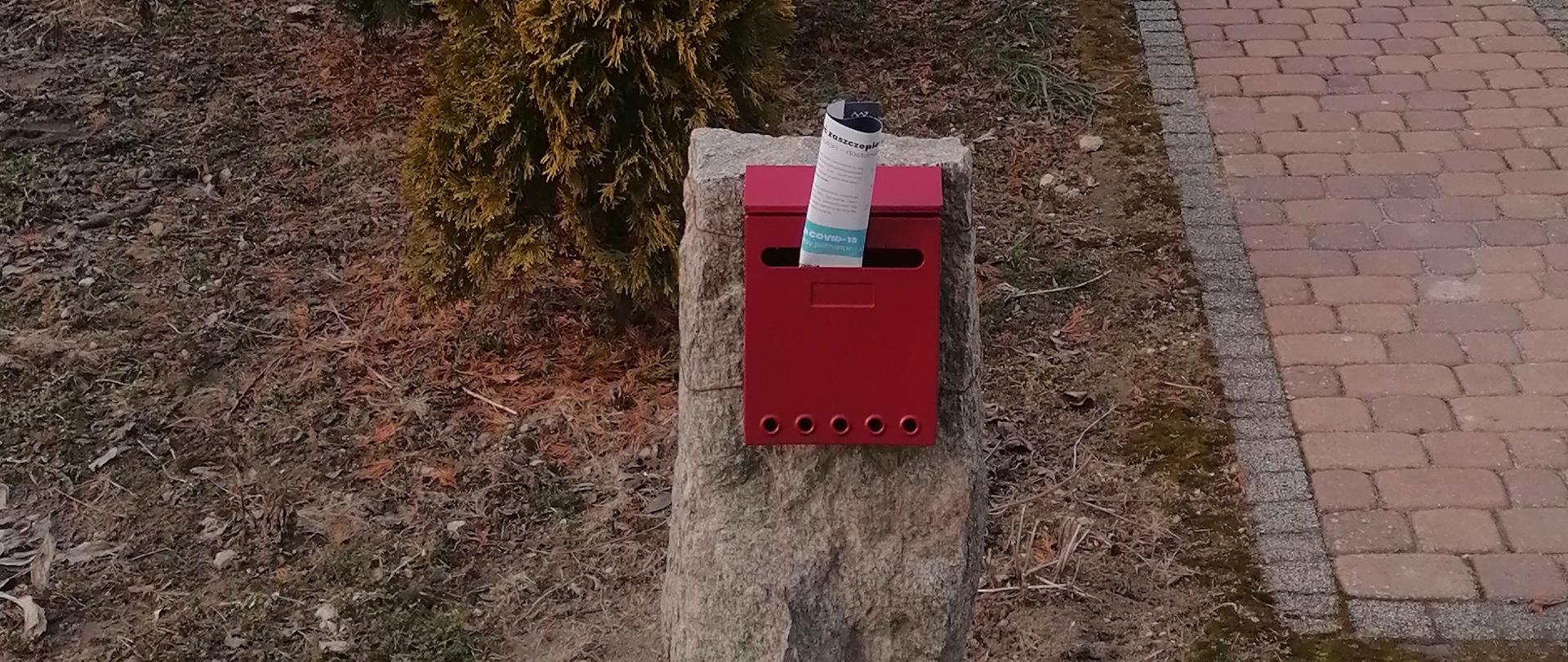 zdjęcie przedstawia zawieszoną na bloku kamiennym skrzynkę na listy do której wsunięto ulotkę dot. szczepień przeciwko Covid 19.