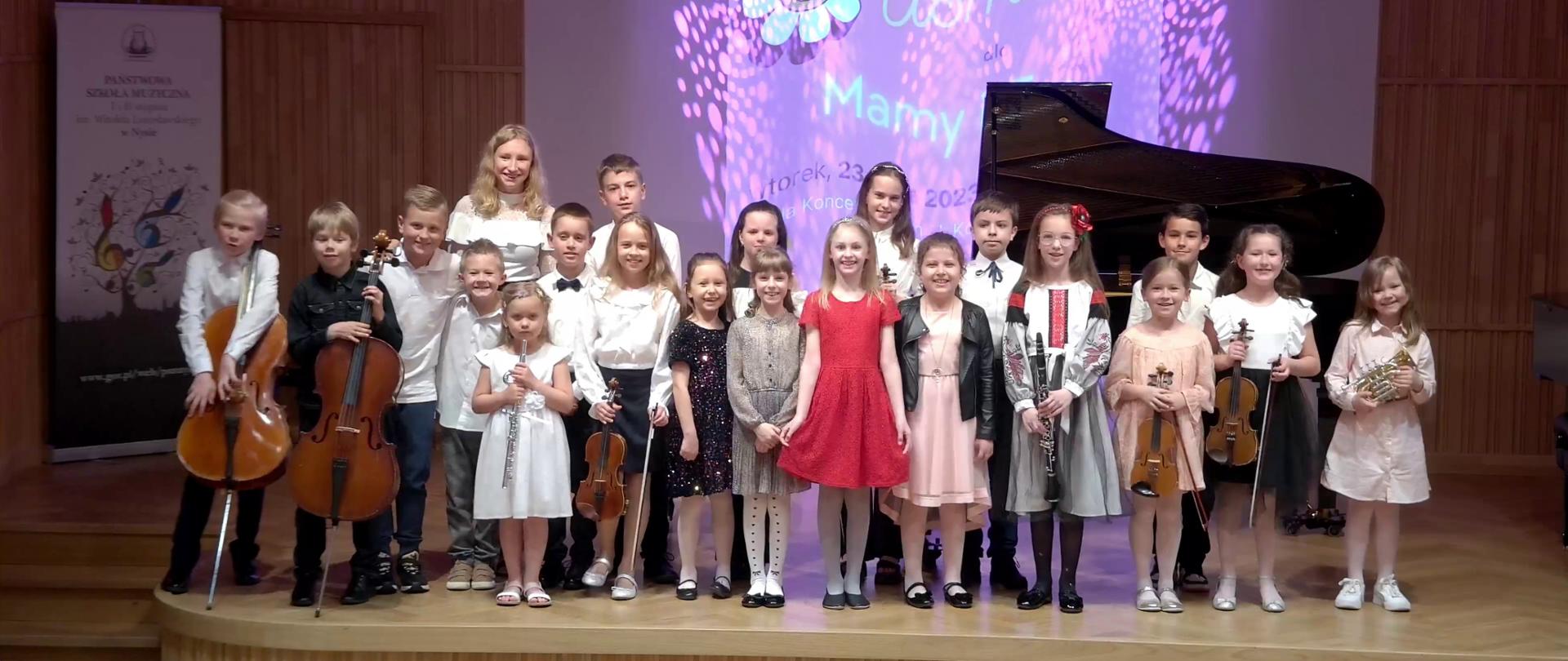 Zdjęcie. na estradzie stoją młodzi wykonawcy koncertu . Część z nich trzyma swoje instrumenty. Za nimi stoi czarny fortepian. Uśmiechają się. W tle wyświetlony jest baner "Uśmiech dla mamy i taty".