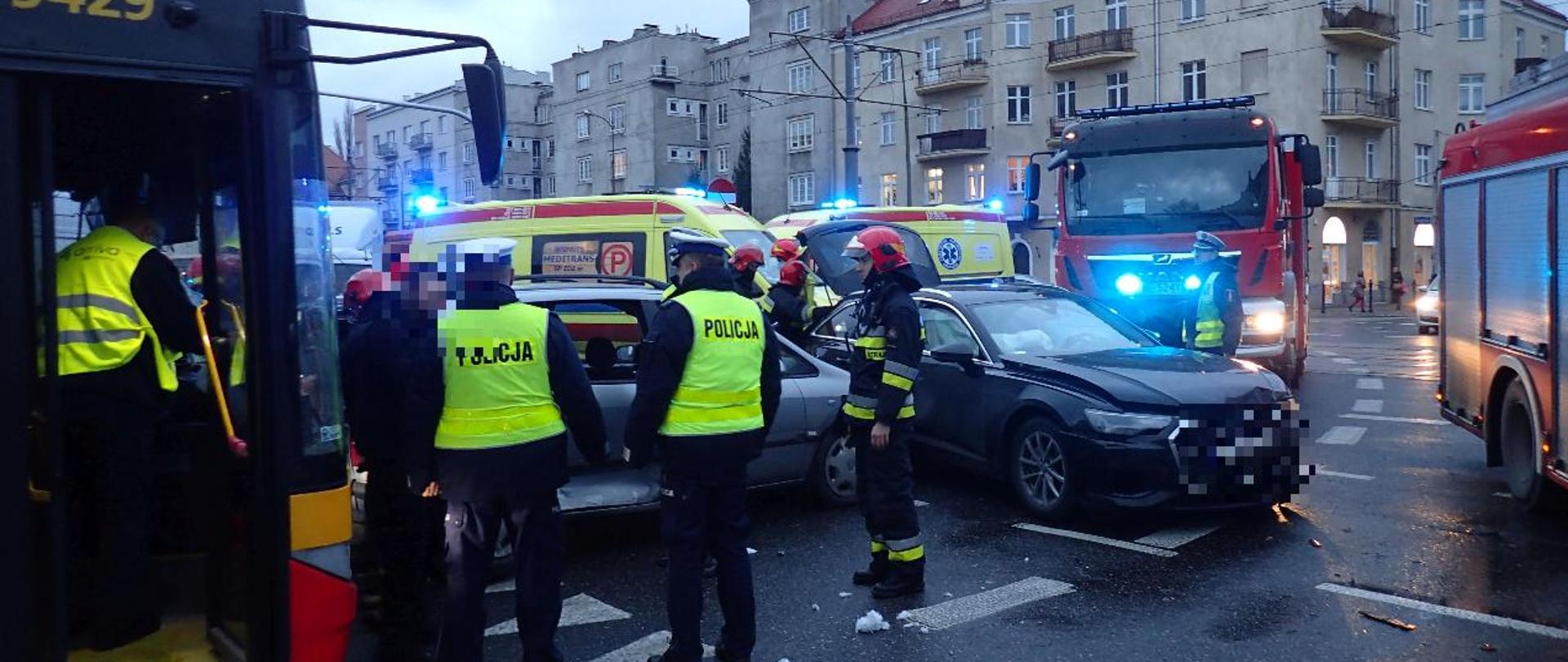 na zdjęciu widać dwa rozbite samochody osobowe oraz uszkodzony autobus komunikacji miejskiej. W tle dwie karetki pogotowia oraz wozy strażackie.