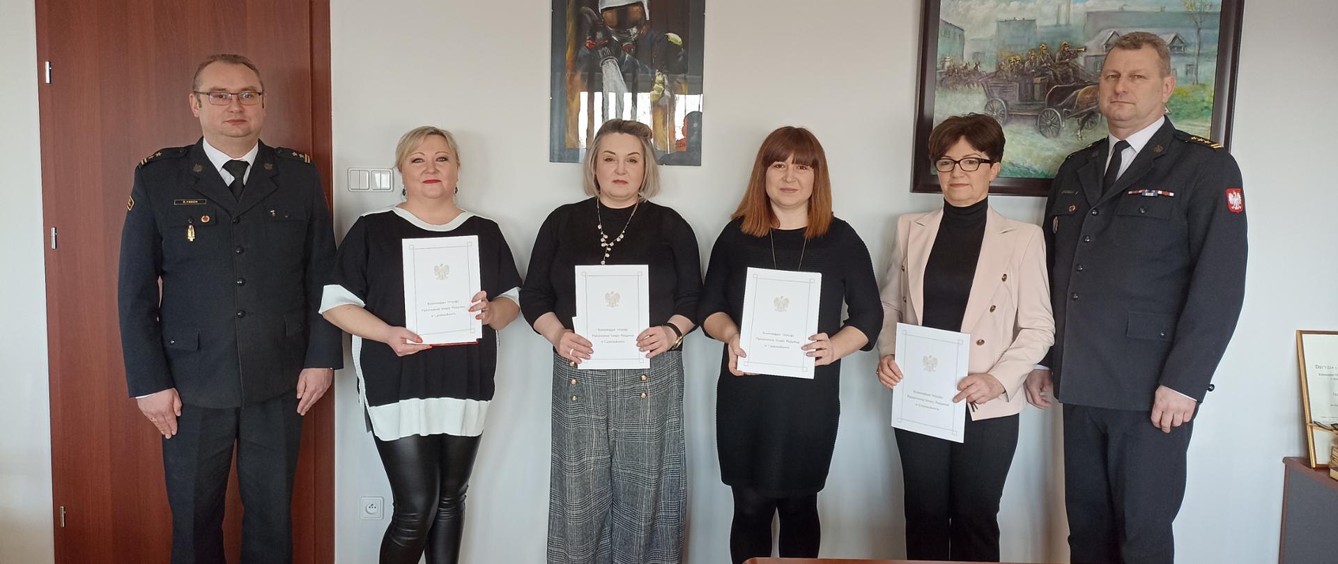 Na zdjęciu wewnątrz pomieszczenia do zdjęcia ustawione sześć osób dwóch funkcjonariuszy Państwowej Straży Pożarnej oraz cztery kobiety. Kobiety trzymają przed sobą teczki na dokumenty z umieszczonym na nich orłem i napisem Komenda Miejska Państwowej Straży Pożarnej w Częstochowie