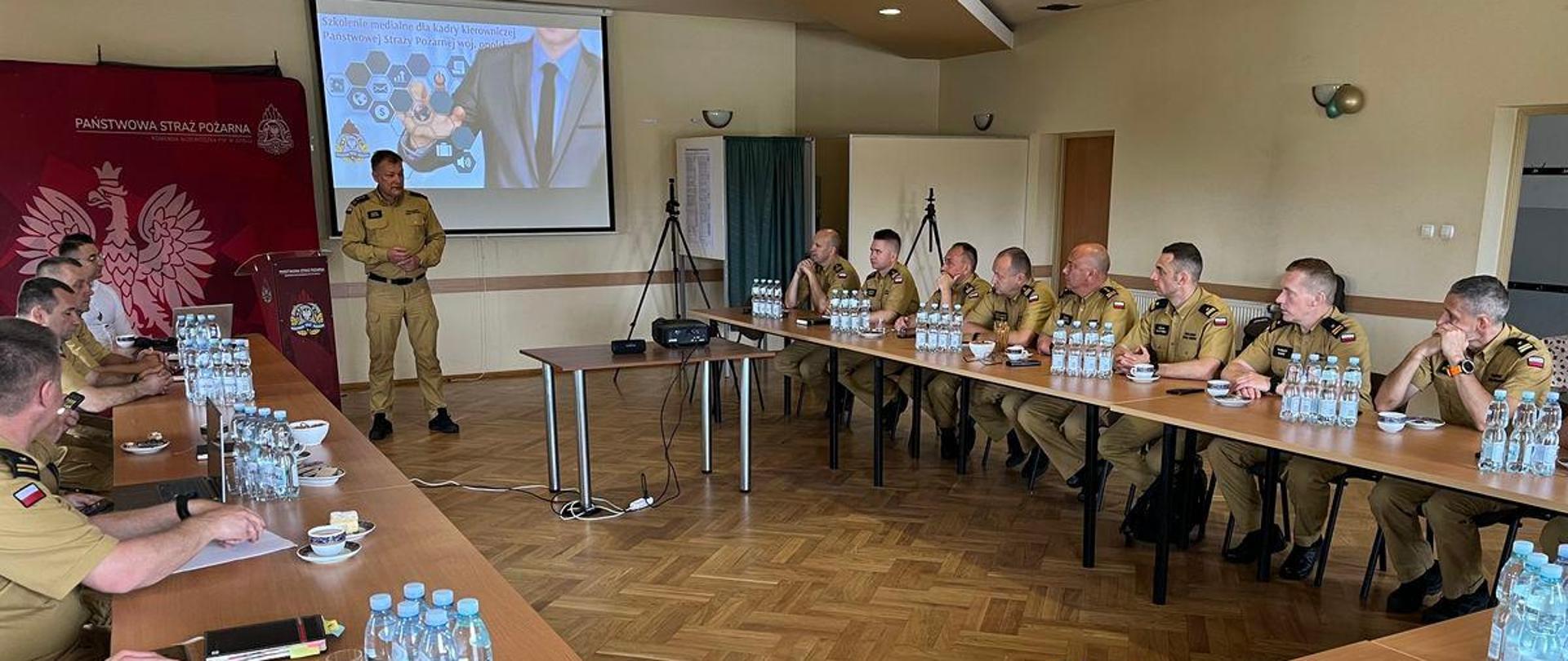 Funkcjonariusze Państwowej Straży Pożarnej siedzą przy stołach w umundurowaniu służbowym słuchając wystąpienia Opolskiego Komendanta Wojewódzkiego Państwowej Straży Pożarnej rozpoczynającego warsztaty. Strażacy zgromadzeni na sali w jednostce OSP Opole - Bierkowice.