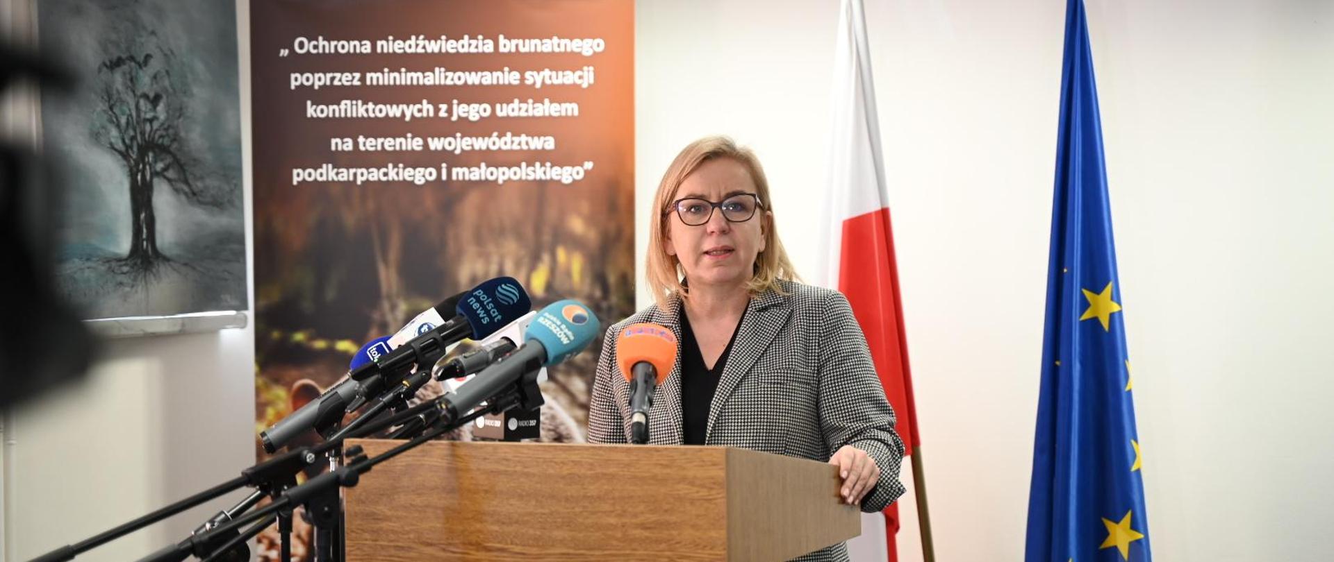 Konferencja ministry klimatu i środowiska Pauliny Hennig-Kloski i przedstawicieli NFOŚiGW oraz RDOŚ w Rzeszowie na temat ochrony niedźwiedzia brunatnego i poprawy bezpieczeństwa mieszkańców Podkarpacia i Małopolski. Na zdjęciu Pani Minister/ kobieta w garniturze w kostiumie drobną kratkę stoi za drewnianą mówicą i przemawia. Za nią flagi polski i UE oraz baner w brązowym kolorze z nazwą programu, który dofinansuje poprawę bezpieczeństwa mieszkańców. 