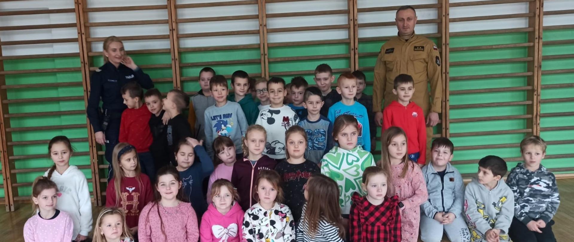 Na zdjęciu znajdują się dzieci siedzące na podłodze oraz strażak i policjantka