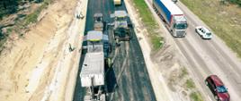 Fotografia lotnicza obrazująca układanie podbudowy drogi z mieszanki mineralno-asfaltowej. Zdjęcie obrazuje odcinek dwujezdniowej drogi. Na jezdni po prawej stronie odbywa się ruch dwukierunkowy, na jezdni lewej trwają pracę asfaltowe. Układanie podbudowy odbywa się przy użyciu dwóch rozściełaczy, pojazdów ciężarowych dowożących gorącą mieszankę, ora czterech walców zagęszczających.