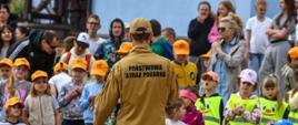 Strażak z Państwowej Straży Pożarnej, widziany od tyłu, stoi przed grupą dzieci i dorosłych. Dzieci noszą kolorowe ubrania i pomarańczowe czapki, część z nich ma na sobie odblaskowe kamizelki z uśmiechniętymi buźkami.