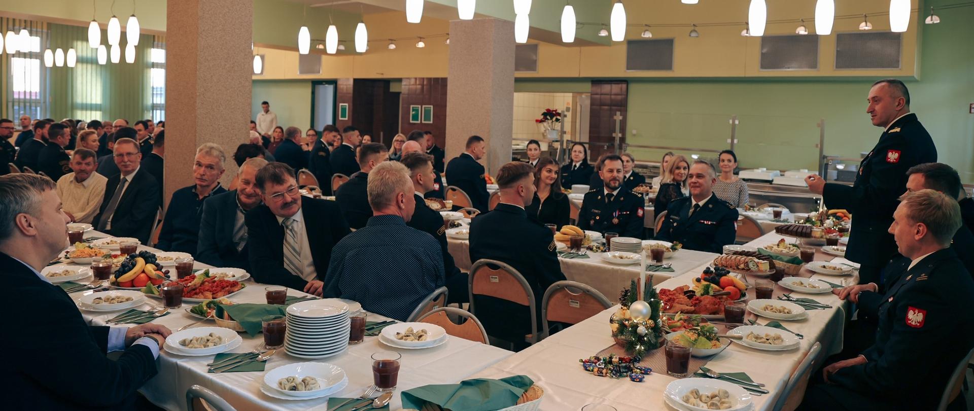 Ujęcie przedstawiające uczestników siedzących przy długim wigilijnym stole. Na pierwszym planie po prawej stronie stoi oficer w mundurze wyjściowym, który przemawia do zgromadzonych. Na stołach widoczne są tradycyjne dania (m.in. pierogi), stroiki świąteczne ze świecami oraz owoce. Sala jest jasna i przestronna.