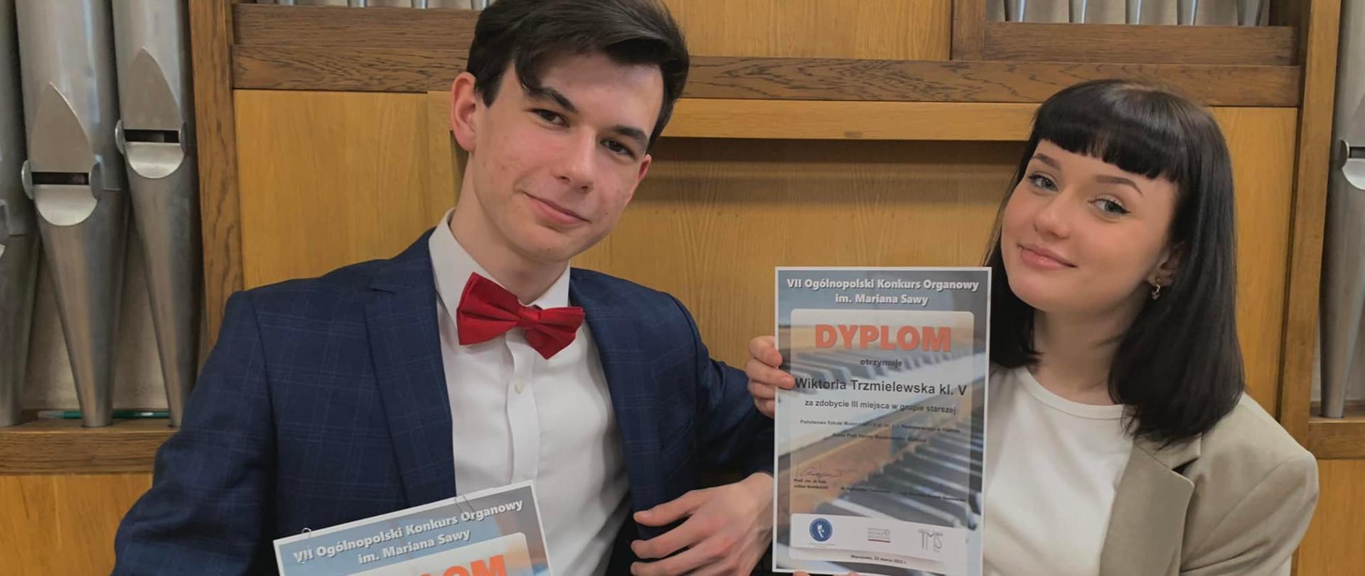 Uśmiechnięty chłopak i uśmiechnięta dziewczyna trzymają dyplomy, siedzą na aławeczce, w tle organy.