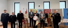 Kompozycja tego zdjęcia j to fotografia grupowa oficjeli w mundurach i strojach wizytowych, ustawionych w rzędzie. Różnica polega na tym, że w centrum stoi inny, młodszy chłopiec. Ubrany jest w elegancką, białą koszulę i ciemne spodnie. Chłopiec dumnie prezentuje do obiektywu otrzymany dyplom w czerwonej ramce i również trzyma w ręku jasną, materiałową torbę. Mężczyzna z mikrofonem z prawej strony jest w trakcie mówienia.
