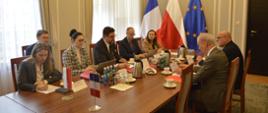 W małej sali przy owalnym stole siedzi siedem osób, za nimi zasłonięte firankami okna, pod ścianą flagi Polski, Francji i UE.