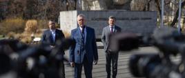 Minister Tomasz Siemoniak stoi przy mikrofonie. Za nim dwóch mężczyzn. W tle pomnik. 