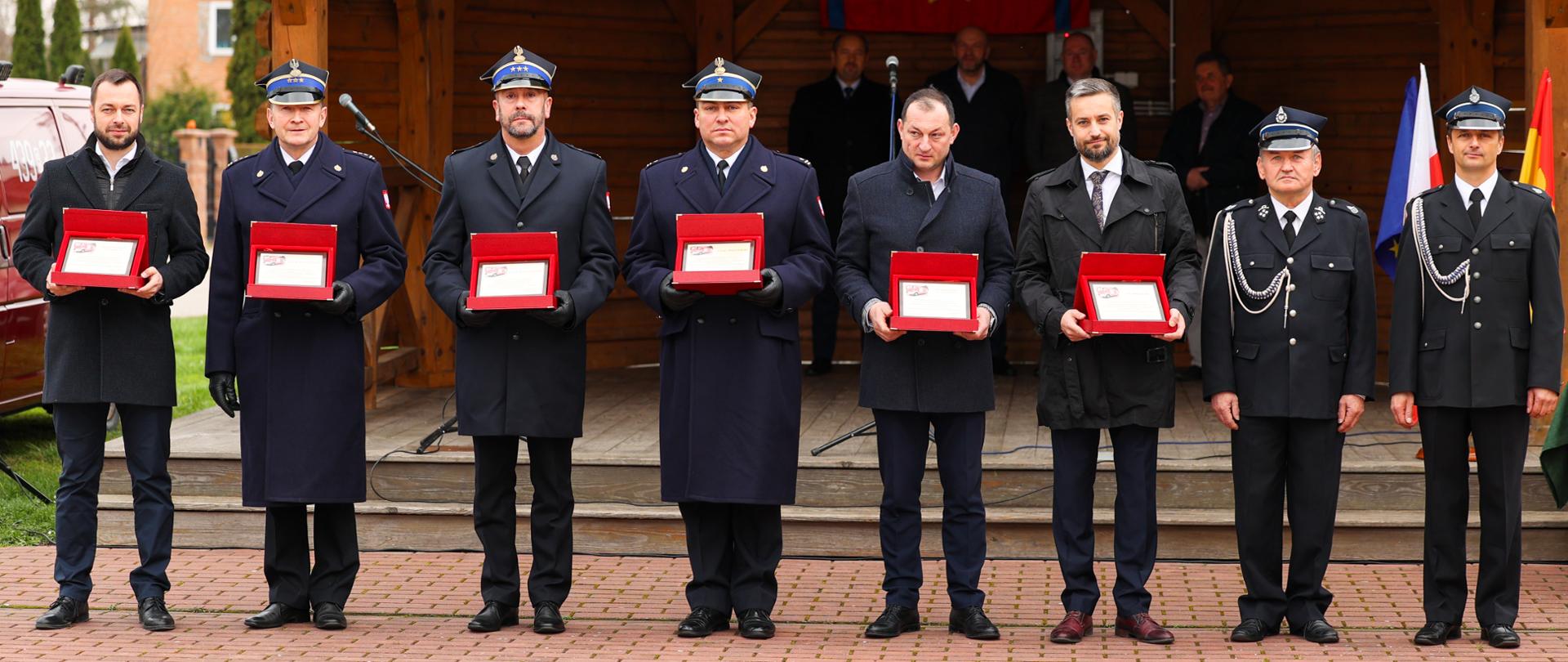 Zdjęcie przedstawia zaproszonych gości trzymających w dłoniach pamiątkowe tablice. Strażacy ubrani są w umundurowanie wyjściowe.