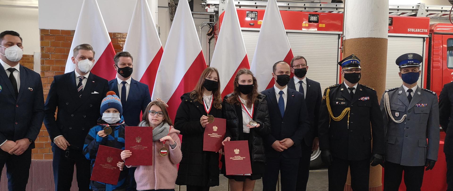 Na pierwszym planie dzieci, które otrzymały medale "Młody Bohater" od lewej Marcin Kochański, Nadia Pyszna, Zofia Degórska, Martyna Góralska. Po prawej stronie wicewojewoda Radosław Kempiński, Kujawsko-Pomorski Komendant Wojewódzki Państwowej Straży Pożarnej st. bryg. Jacek Kaczmarek, Zastępca Komendanta Wojewódzkiego Policji w Bydgoszczy insp. Mirosław Elszkowski, Burmistrz Kruszwicy Dariusz Witczak. Po lewej stronie Prezes Zarządu Wodnego Ochotniczego Pogotowia Ratunkowego Województwa Kujawsko-Pomorskiego Maciej Banachowski, Dyrektor Wydziału Bezpieczeństwa i Zarządzania Kryzysowego Kujawsko-Pomorskiego Urzędu Wojewódzkiego w Bydgoszczy Rafał Rewoliński, W tle flagi państwowe i wóz pożarniczy
