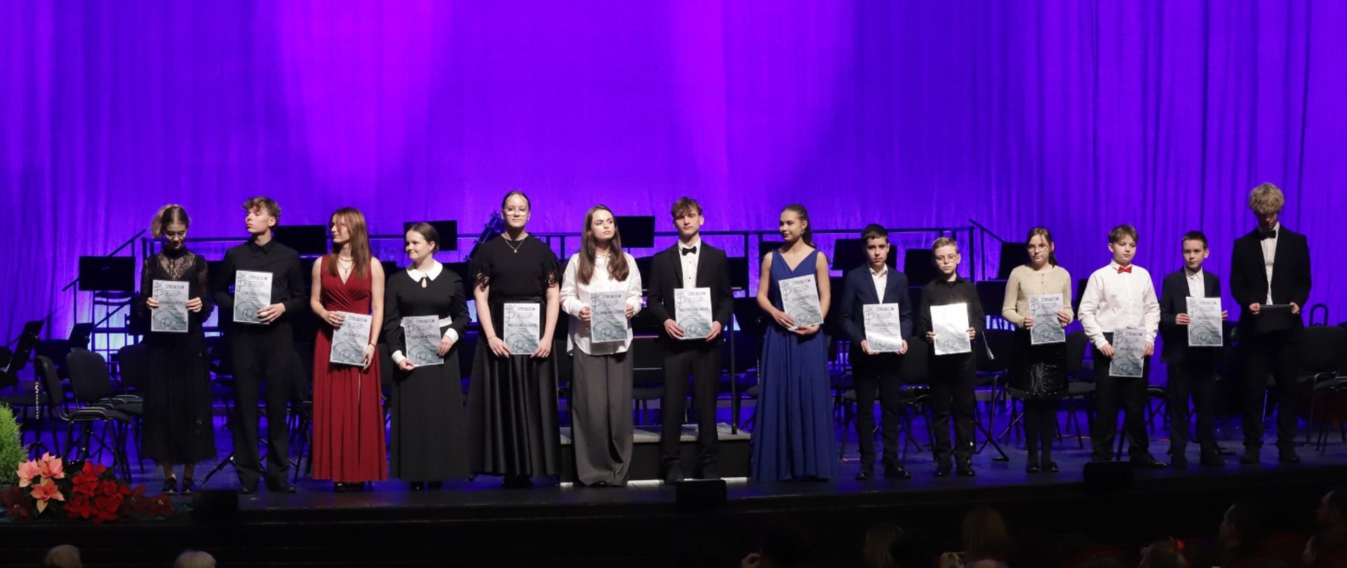 Na zdjęciu widać 14 młodych osób stojących na scenie Teatru Ziemi Rybnickiej i ustawionych w jednym rzędzie. Każda z nich trzyma w rękach dyplom. Osoby są elegancko ubrane — dziewczęta w długich sukniach, a chłopcy w garniturach lub koszulach.
Tło sceny jest oświetlone fioletowym światłem, a za nimi widać ustawione pulpity i instrumenty. Na pierwszym planie, w dolnym rogu, widać fragment dekoracji z kwiatami. Całość to uroczystość wręczenia stypendiów szkolnych i listów gratulacyjnych dyrektora za otrzymanie stypendium MKiDN.