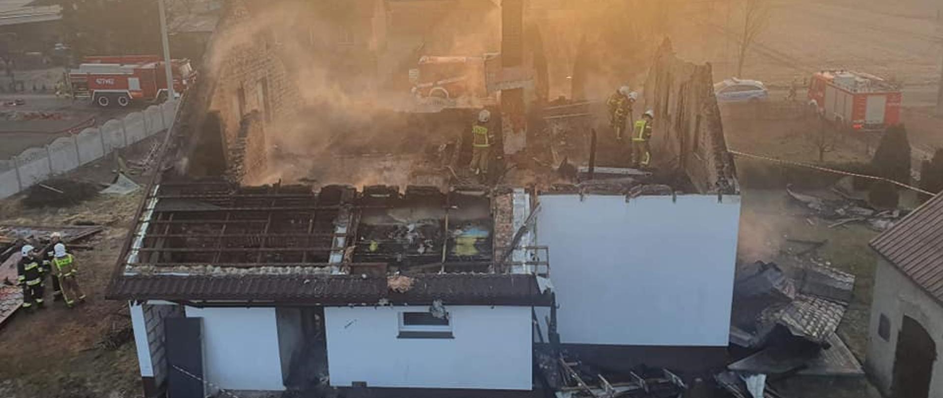 Widok z kosza drabiny mechanicznej. Biały budynek bez dachu. Strażacy dogaszają ogniska pożaru na poddaszu.