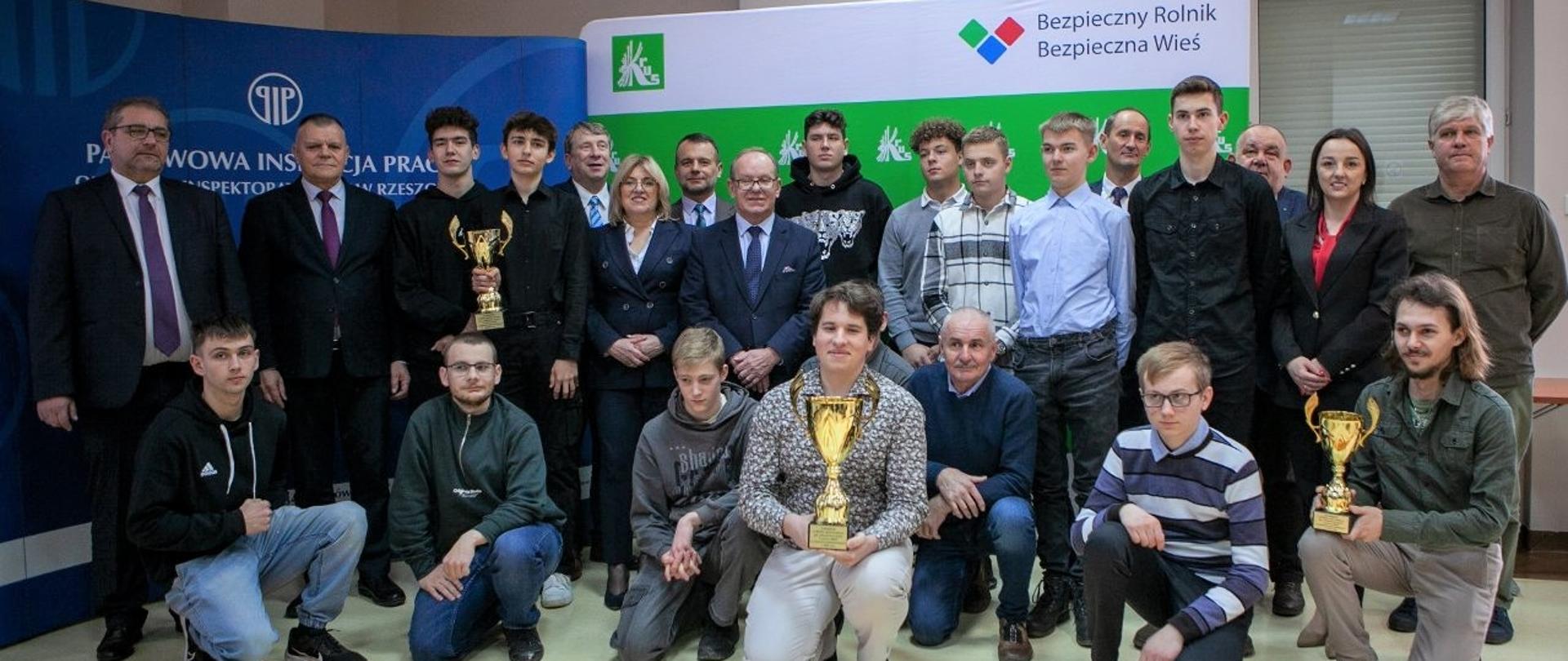 Na tle ścianki KRUS oraz PIP Laureaci Olimpiady (trzymając zdobyte puchary) wraz
z przedstawicielami Organizatorów pozują do zdjęcia.
